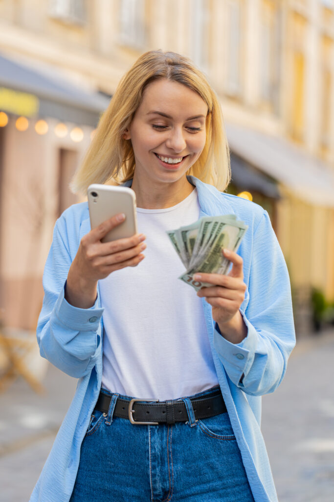 happy blonde woman counting money dollar cash, use smartphone calculator app on urban city street
